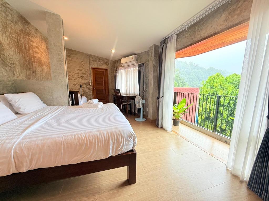 ห้องพักเชียงใหม่สไตล์โมเดิร์น ประกอบด้วยเตียงสีขาว โต๊ะเล็กและเก้าอี้ พัดลมไฟฟ้า และระเบียงพร้อมวิวต้นไม้สีเขียวและภูเขาไกล.