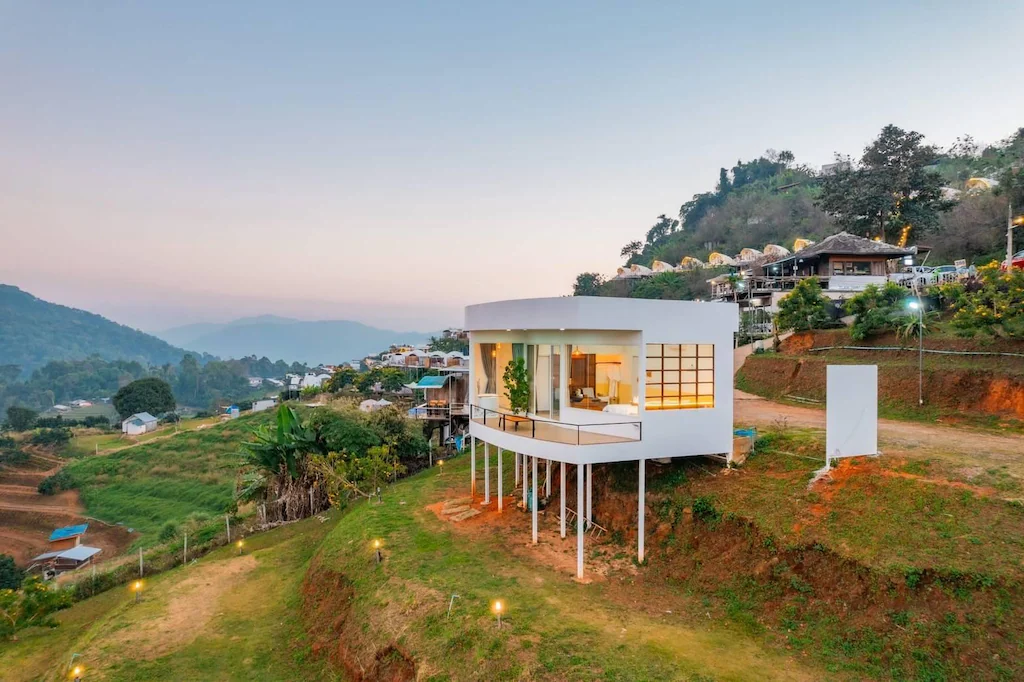 บ้านสีขาวสมัยใหม่บนเสาสูงตั้งอยู่บนเนินหญ้าที่มีภูเขาและอาคารอื่นๆ เป็นฉากหลังในยามพระอาทิตย์ตกดิน ดูคล้ายรีสอร์ทเชียงใหม่ที่มีสไตล์ เหมาะสำหรับการพักผ่อนอย่างสงบ.