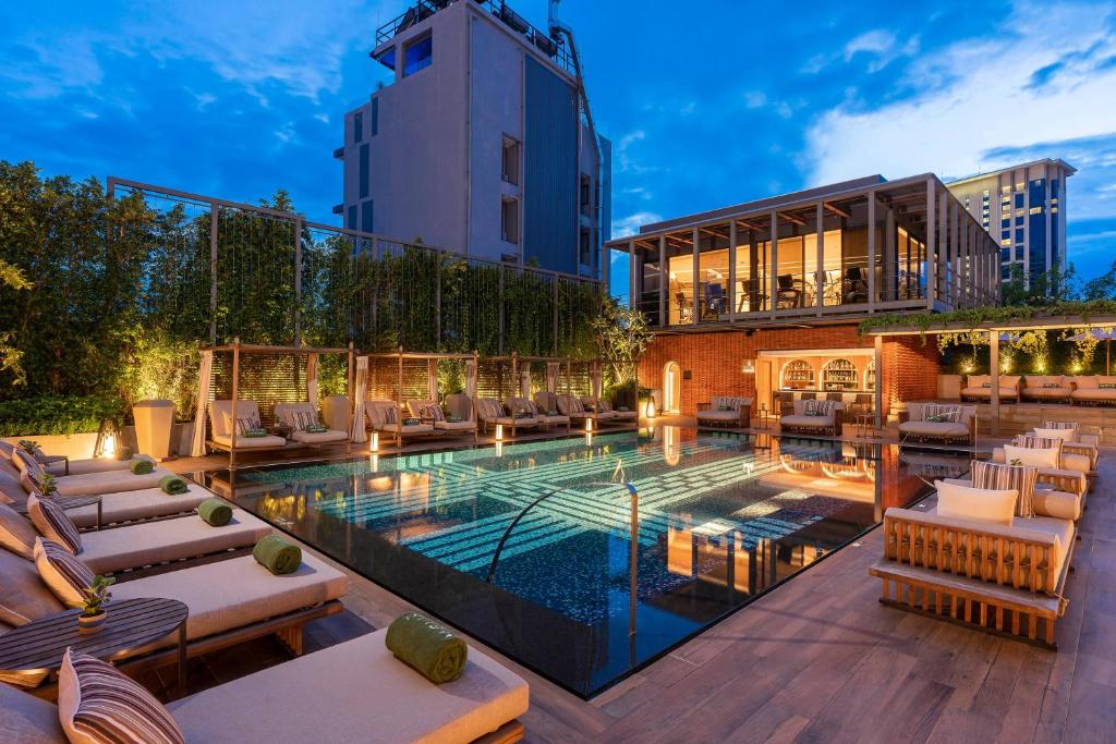 สระว่ายน้ำบนดาดฟ้าล้อมรอบด้วยเก้าอี้อาบแดดและต้นไม้เขียวขจี พร้อมบาร์ทันสมัยและพื้นที่นั่งเล่นในฉากหลังยามเย็น—เหมาะสำหรับการผ่อนคลายที่ โรงแรมเชียงใหม่ ที่มีสไตล์.