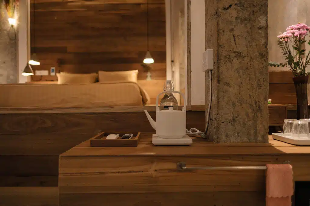 เตียงที่จัดไว้อย่างเรียบร้อยพร้อมผ้าปูที่นอนสีแทนอยู่ด้านหลัง; ด้านหน้าเป็นกาต้มน้ำ แก้วน้ำ และของใช้ในห้องน้ำบนเคาน์เตอร์ไม้ จัดวางอย่างสมบูรณ์แบบสำหรับการพักผ่อนที่ โรงแรมเชียงใหม่ ดอกไม้สีชมพูสดใสในแจกันเพิ่มความสดชื่นอยู่ทางขวา.