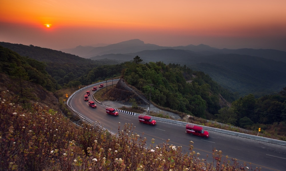 รถสีแดงวิ่งเรียงแถวไปตามถนนภูเขาโค้งในยามพระอาทิตย์ตกดิน โดยมีภูเขาที่ปกคลุมไปด้วยป่าและภูเขาไกลๆ เป็นฉากหลัง—ทัศนียภาพที่สมบูรณ์แบบสำหรับการค้นหา รีสอร์ทเชียงใหม่ ที่เหมาะสำหรับคุณท่ามกลางธรรมชาติ.