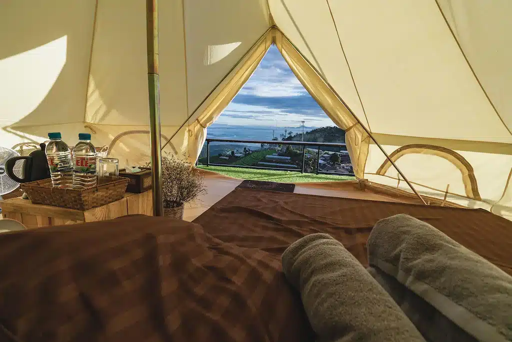 วิวจากภายในเต็นท์ที่รีสอร์ทเชียงใหม่ ประกอบด้วยเตียงนอนที่แสนสบายพร้อมผ้าเช็ดตัว ตะกร้าที่บรรจุขวดน้ำ และผ้าใบเปิดที่เผยให้เห็นเนินเขาที่ปกคลุมไปด้วยหญ้าและท้องฟ้าที่มีเมฆปกคลุมในทิวทัศน์ที่สวยงามของเชียงใหม่.