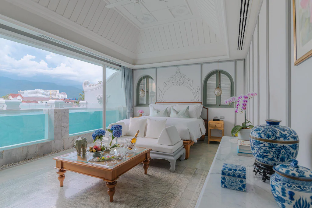 ห้องพักเชียงใหม่ที่สว่างไสวและหรูหรา พร้อมเตียงขนาดใหญ่ ตกแต่งด้วยสีน้ำเงินและขาว มีมุมนั่งเล่นพร้อมอาหารและเครื่องดื่ม และหน้าต่างระเบียงที่มอบวิวเมืองและภูเขาที่สวยงามอย่างเหลือเชื่อ.