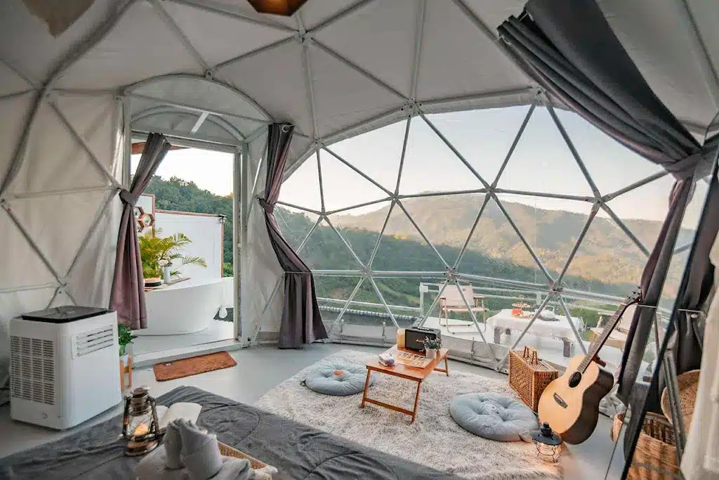 ภายในเต็นท์แกลมปิ้งทรงโดม ณ ที่พักเชียงใหม่ โดดเด่นด้วยหน้าต่างบานใหญ่ที่มองเห็นวิวภูเขา พื้นที่นั่งเล่นแสนสบาย โต๊ะเล็ก กีตาร์ และของตกแต่งแสงไฟอ่อน ๆ—เหมาะอย่างยิ่งสำหรับการพักผ่อนสุดพิเศษใกล้ชิดธรรมชาติ.