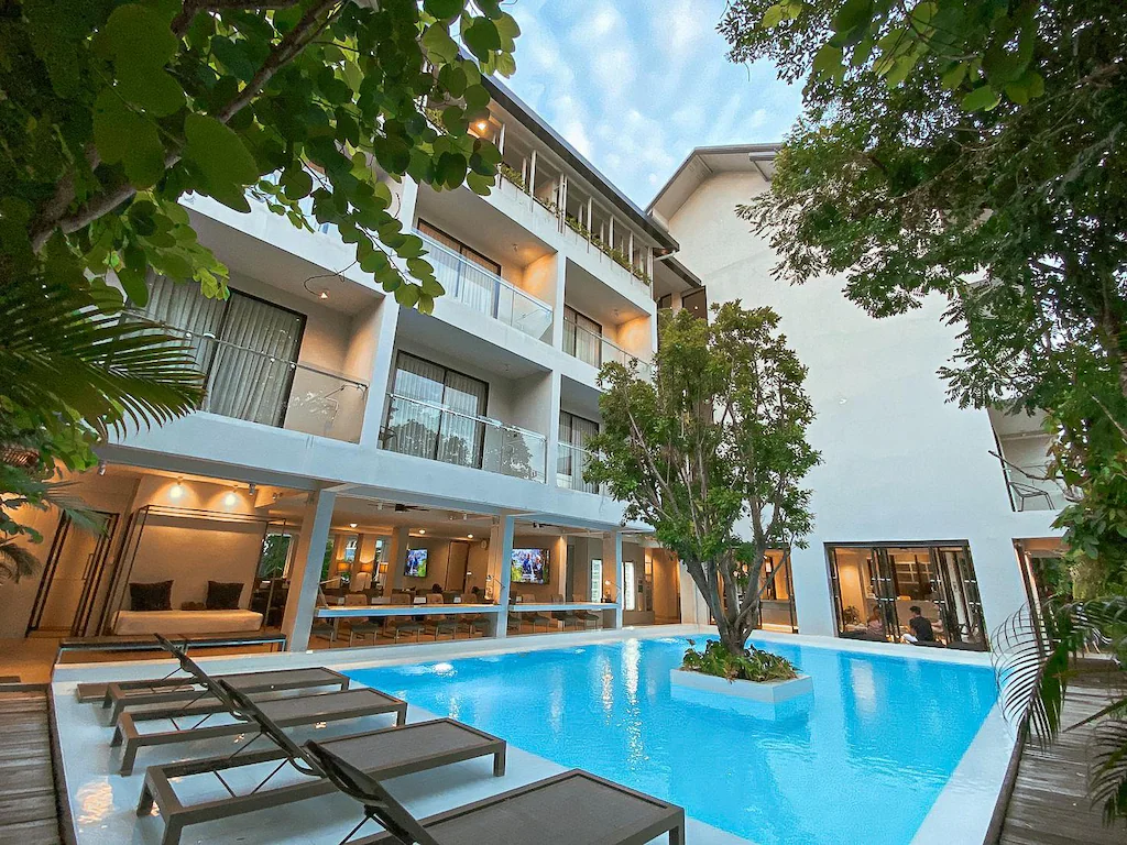 ลานโรงแรมสมัยใหม่พร้อมสระว่ายน้ำรูปสี่เหลี่ยมผืนผ้า เก้าอี้อาบแดด และต้นไม้ ล้อมรอบด้วยอาคารสีขาวสูงสามชั้นที่มีระเบียงและหน้าต่างบานใหญ่—โรงแรมในเชียงใหม่ที่สมบูรณ์แบบสำหรับการเข้าพักครั้งต่อไปของคุณ.