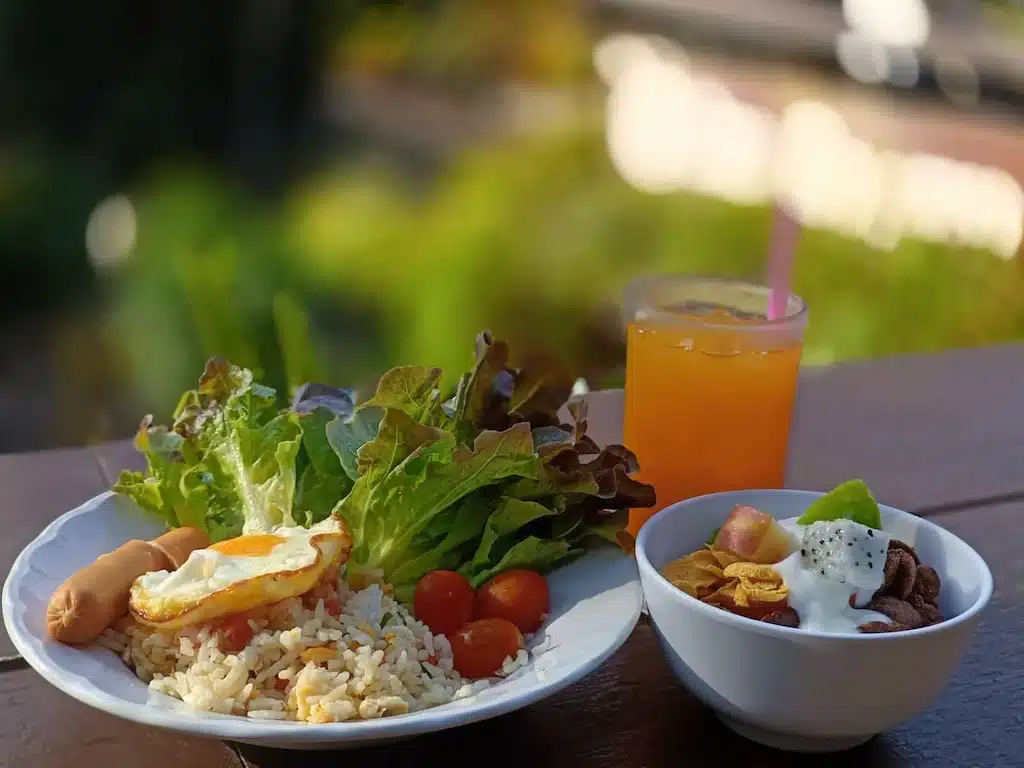 จานอาหารเช้าแสนอร่อยประกอบด้วยข้าว ไข่ดาว ไส้กรอก ผักกาดหอม และมะเขือเทศเชอร์รี่ พร้อมโยเกิร์ตผลไม้และกราโนล่า—เหมาะอย่างยิ่งสำหรับการเริ่มต้นวันใหม่ที่รีสอร์ทเชียงใหม่อันแสนอบอุ่น.