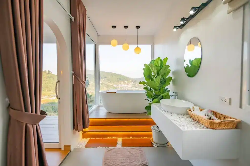 ห้องน้ำสไตล์โมเดิร์นที่โรงแรมเชียงใหม่ พร้อมอ่างอาบน้ำแบบลอยตัวติดหน้าต่างบานใหญ่ อ่างล้างหน้าแบบลอยตัว กระจกกลม ต้นไม้ในกระถาง และแสงไฟอบอุ่น—เหมาะสำหรับการผ่อนคลายพร้อมชมวิวทิวทัศน์ภายนอกที่สวยงาม.