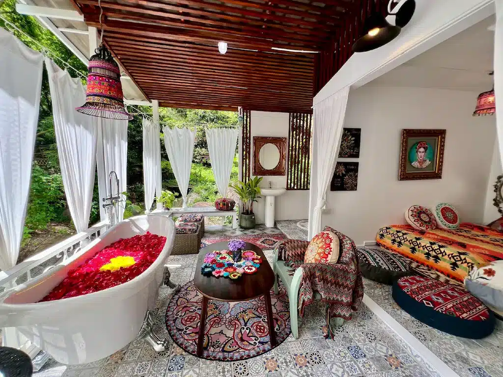 ห้องน้ำกลางแจ้งสไตล์ผสมผสานที่ ที่พักเชียงใหม่ พร้อมอ่างอาบน้ำขาสิงห์ที่เต็มไปด้วยกลีบกุหลาบ พรมลายสวย เฟอร์นิเจอร์โบฮีเมียน ผ้าหลากสีสัน และม่านสีขาวที่เปิดออกสู่พื้นที่สีเขียวชอุ่ม.
