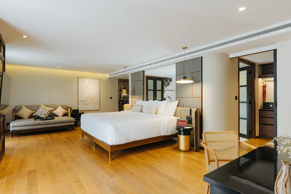 ห้องพักโรงแรมสมัยใหม่พร้อมเตียงขนาดใหญ่ โซฟา โต๊ะทำงาน และเก้าอี้บนพื้นไม้ มีกระจกบานใหญ่ แสงไฟนุ่มนวล และพื้นที่ตู้เสื้อผ้าแบบเปิดโล่งอยู่ด้านหลัง—เหมาะอย่างยิ่งสำหรับการเข้าพักที่โรงแรมเชียงใหม่หรือรีสอร์ทเชียงใหม่ของคุณ.