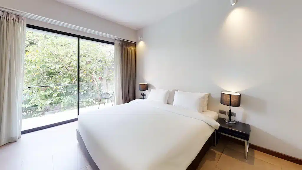ห้องนอนสมัยใหม่พร้อมเตียงขนาดใหญ่ โต๊ะข้างเตียงสองตัวพร้อมโคมไฟ และประตูบานเลื่อนกระจกที่เปิดออกสู่ระเบียงพร้อมต้นไม้สีเขียวด้านนอก—เหมาะสำหรับผู้ที่ต้องการที่พักเชียงใหม่ที่อบอุ่นท่ามกลางธรรมชาติ.