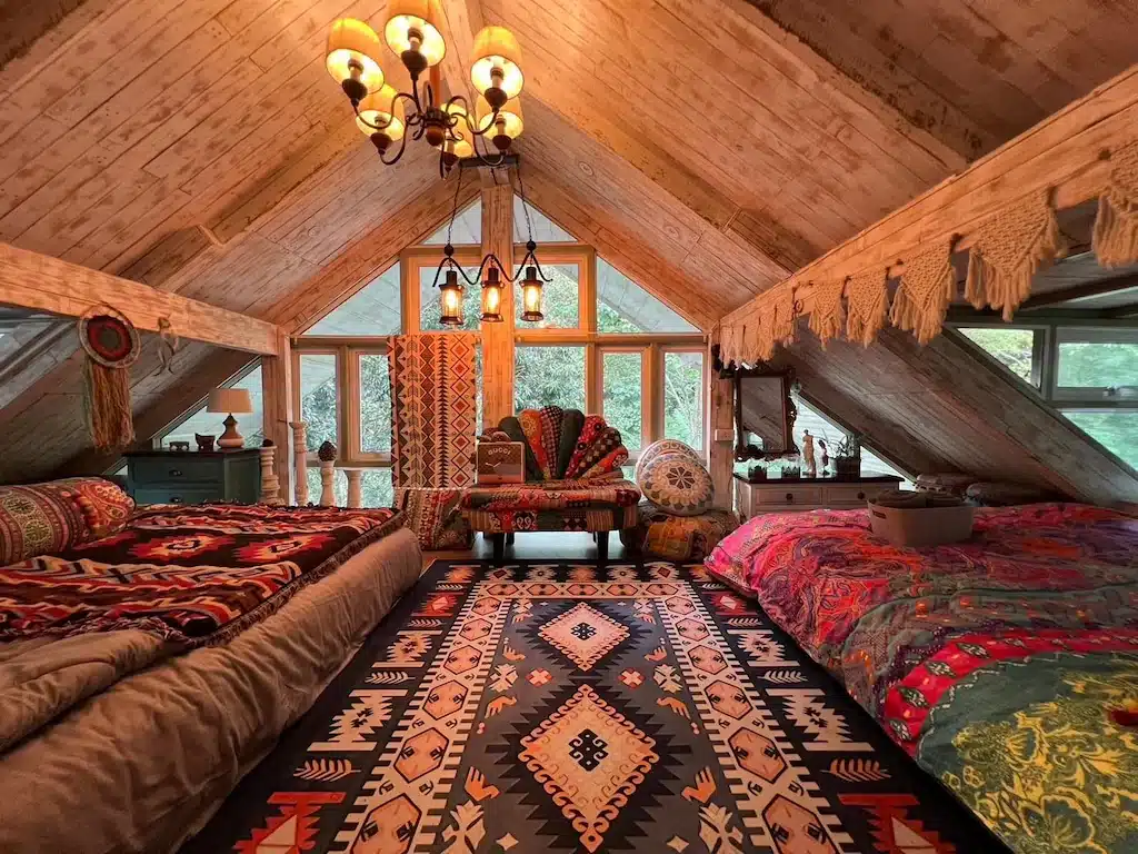 ห้องนอนใต้หลังคาที่แสนอบอุ่นในโรงแรมเชียงใหม่ พร้อมเตียงสองเตียง, เก้าอี้โซฟา, พรมลาย, ผ้าสีสันสดใส, ผนังไม้, และหน้าต่างบานใหญ่ที่ให้แสงสว่างจากธรรมชาติเข้ามา.