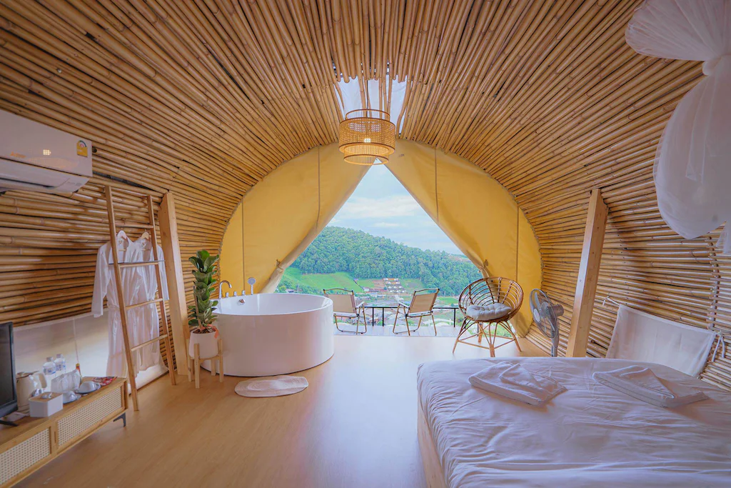 ห้องทรงโดมที่อบอุ่นในรีสอร์ทเชียงใหม่ ผนังไม้ไผ่ เตียงนอน อ่างอาบน้ำ เก้าอี้ และวิวทิวทัศน์ของภูเขาเขียวขจีผ่านหน้าต่างบานใหญ่ที่เปิดโล่ง.