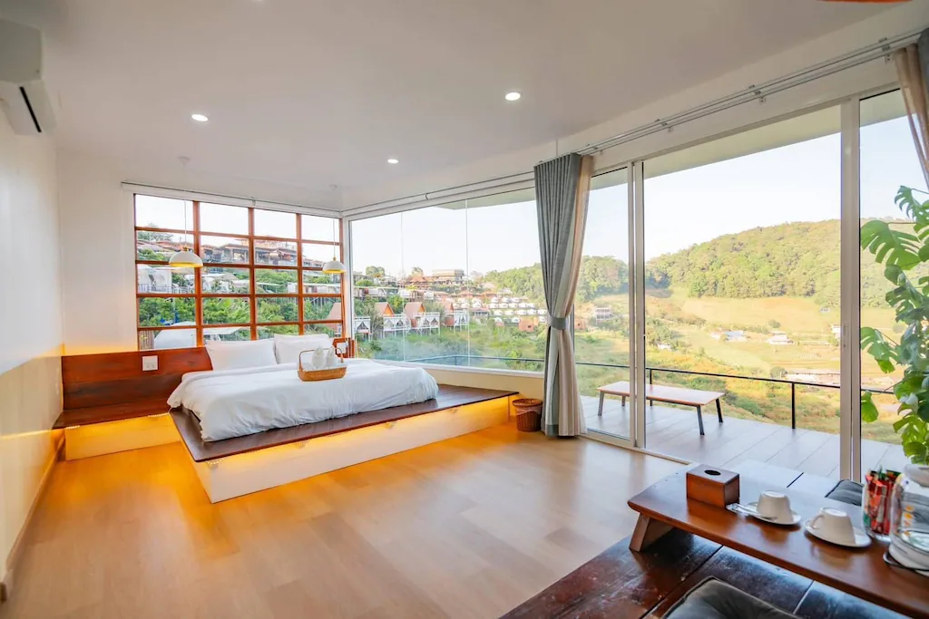 ห้องนอนสไตล์โมเดิร์นที่ ที่พักเชียงใหม่ พร้อมเตียงแพลตฟอร์ม หน้าต่างบานใหญ่ และวิวภูเขาที่สวยงาม การตกแต่งแบบมินิมอลประกอบด้วยโต๊ะเล็ก ๆ พร้อมชุดน้ำชา และผ้าม่านที่เปิดไว้ด้านหนึ่ง.