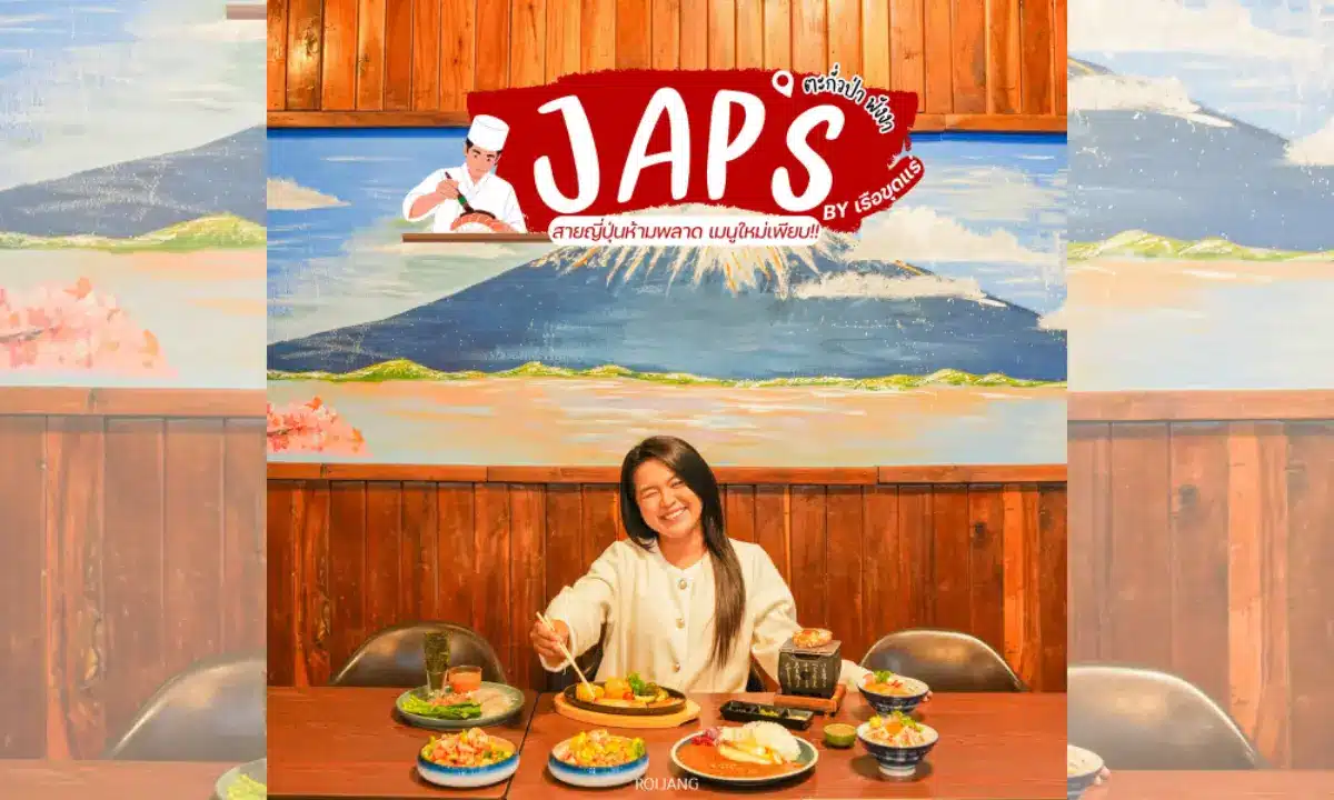 ผู้หญิงคนหนึ่งนั่งอยู่ที่โต๊ะซึ่งมีอาหารญี่ปุ่นหลากหลายชนิดวางอยู่ตรงหน้าภาพจิตรกรรมฝาผนังของภูเขาฟูจิ ภายใต้ป้ายชื่อร้านอาหารที่เขียนว่า "JAPs ตะกั่วป่า.