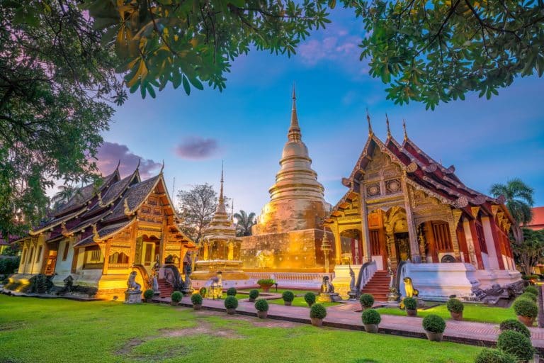 เจดีย์ทองคำและอาคารวัดที่ประดับประดาอย่างวิจิตรที่วัดพระสิงห์ หนึ่งในสถานที่ท่อง เที่ยวเชียงใหม่ ยอดนิยม ล้อมรอบด้วยต้นไม้เขียวขจีและสนามหญ้าภายใต้ท้องฟ้าสีสันสดใสยามพระอาทิตย์ตกดิน.