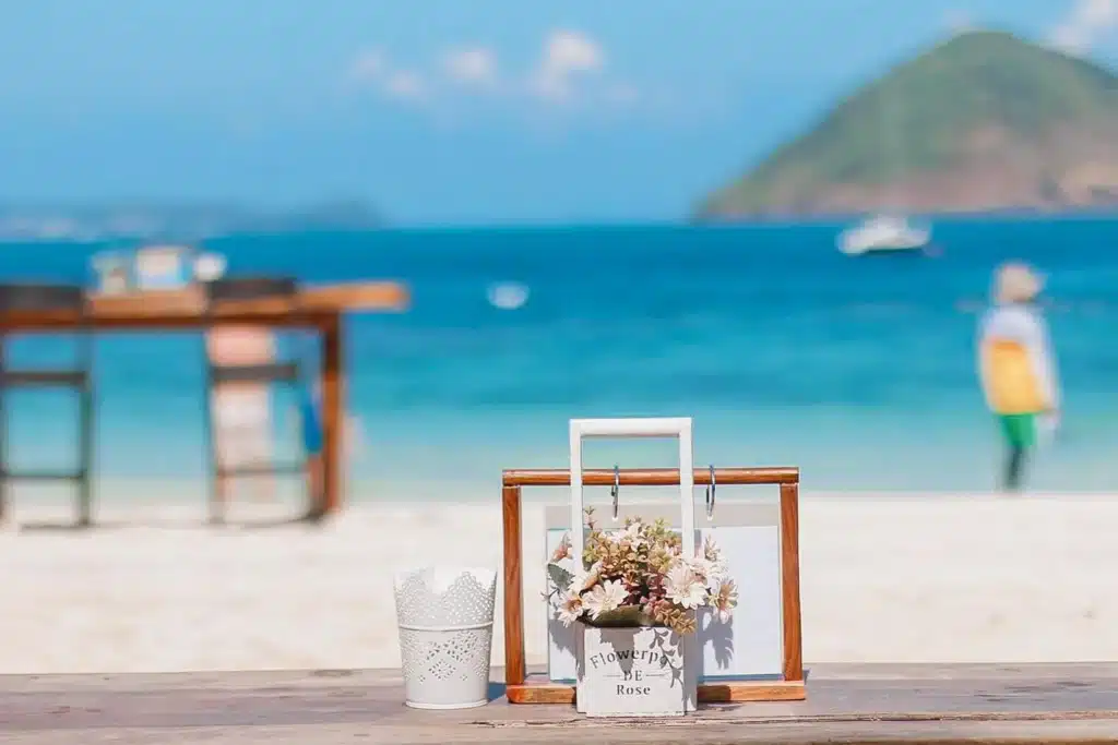 การจัดดอกไม้ขนาดเล็กและภาชนะสีขาววางอยู่บนโต๊ะไม้ที่มองเห็นชายหาดทรายพร้อมทะเลสีฟ้า—สถานที่ที่เหมาะสำหรับใครก็ตามที่กำลังมองหา ที่เที่ยวใกล้กรุงเทพ และทิวทัศน์ธรรมชาติที่เงียบสงบ.