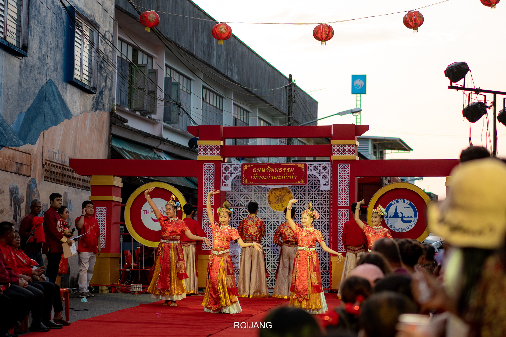 กลุ่มนักเต้นในชุดประจำชาติแสดงบนเวทีพรมแดงกลางแจ้ง โดยมีผู้ชมชมการแสดงและโคมไฟสีแดงแขวนอยู่ด้านบน—เป็นภาพที่น่าหลงใหลสำหรับผู้ที่ต้องการ เที่ยวใกล้กรุงเทพ.