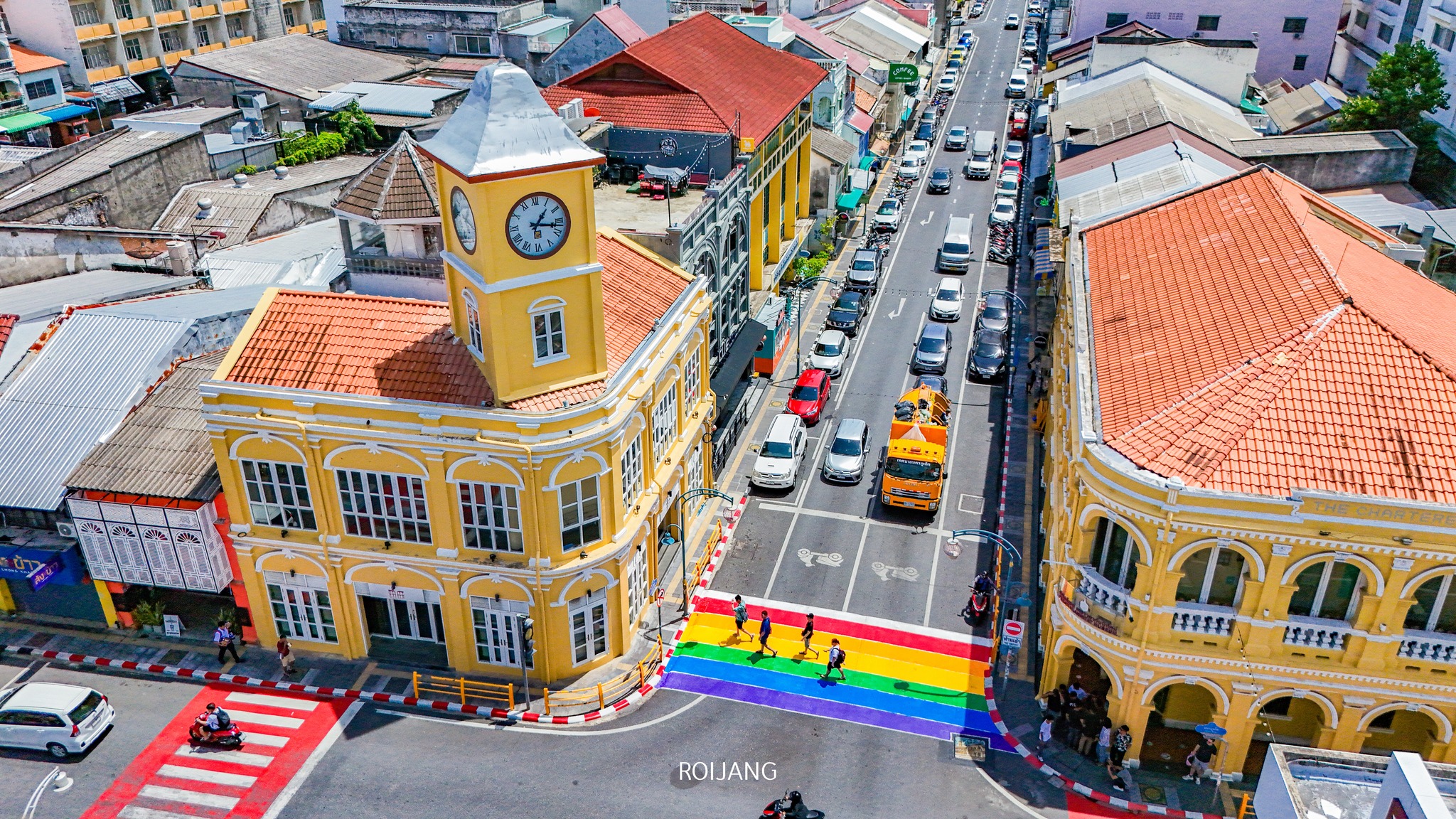 ภาพมุมสูงของสี่แยกในเมืองที่มีอาคารสไตล์โคโลเนียลสีเหลืองและทางม้าลายสีรุ้ง ซึ่งเต็มไปด้วยรถยนต์และคนเดินเท้า—เป็นภาพที่คึกคักในหนึ่งในสถานที่ท่องเที่ยวใกล้กรุงเทพฯ.