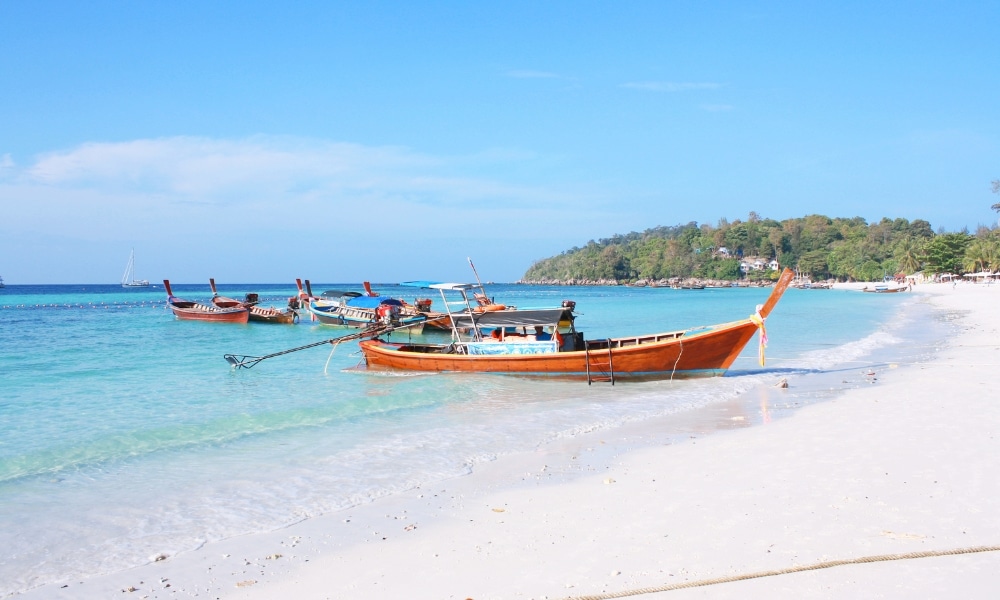 เรือไม้จอดทอดสมออยู่ในน้ำตื้นใสสีฟ้า ใกล้ชายหาดทรายขาวที่มีต้นไม้เขียวขจีอยู่ใต้ท้องฟ้าแจ่มใส—ภาพที่สมบูรณ์แบบสำหรับผู้ที่มองหาสถานที่ท่องเที่ยวธรรมชาติใกล้กรุงเทพฯ เพื่อพักผ่อนและคลายความเครียด.