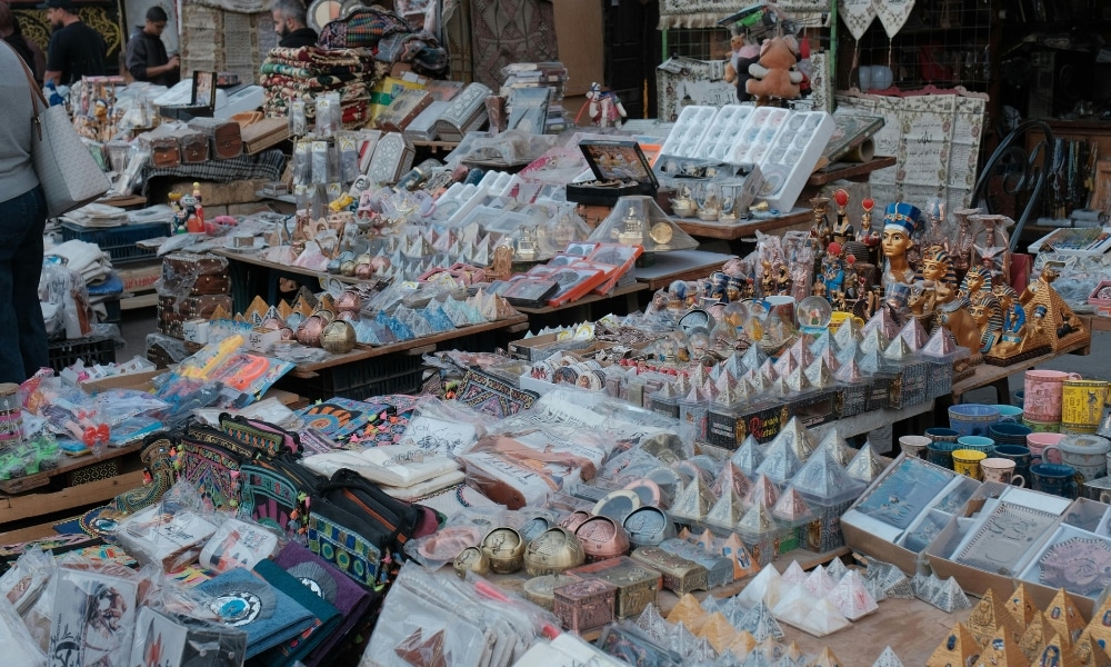 แผงขายของในตลาดจัดแสดงของที่ระลึกหลากหลายชนิด เหมาะสำหรับผู้ที่ต้องการเที่ยวใกล้กรุงเทพฯ รวมถึงพีระมิดจำลอง รูปปั้นขนาดเล็ก โปสการ์ด และถุงผ้าสีสันสดใสที่จัดเรียงอย่างเรียบร้อยบนโต๊ะ.