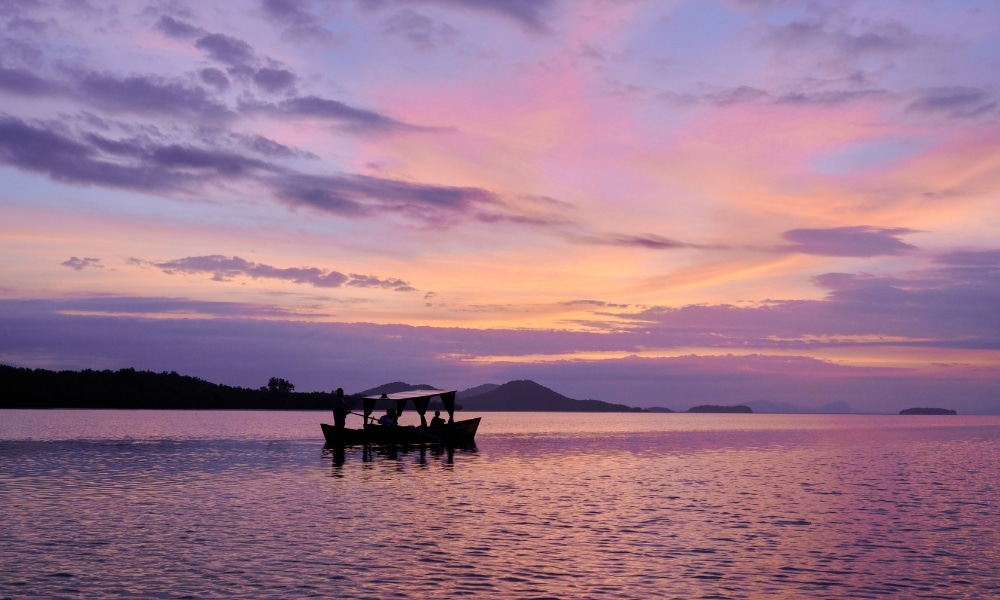 เรือลำเล็กที่มีผู้คนหลายคนกำลังลอยอยู่เป็นเงาดำตัดกับท้องฟ้ายามพระอาทิตย์ตกที่เต็มไปด้วยสีสันเหนือทะเลสาบที่เงียบสงบ โดยมีภูเขาไกลๆ อยู่บนขอบฟ้า—เหมาะสำหรับผู้ที่กำลังมองหาสถานที่ท่องเที่ยวธรรมชาติใกล้กรุงเทพฯ ในช่วงวันหยุดพักผ่อน.