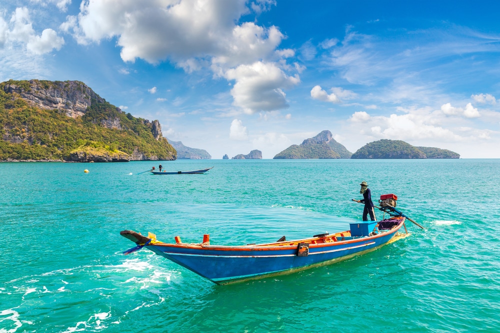 บุคคลหนึ่งยืนอยู่บนเรือไม้สีน้ำเงินในน้ำสีฟ้าคราม มีเกาะหินและเรือลำอื่นปรากฏอยู่ภายใต้ท้องฟ้าที่มีเมฆบางส่วน—เหมาะสำหรับการเที่ยวใกล้กรุงเทพฯ หรือผู้ที่มองหาสถานที่ท่องเที่ยวธรรมชาติใกล้กรุงเทพฯ.