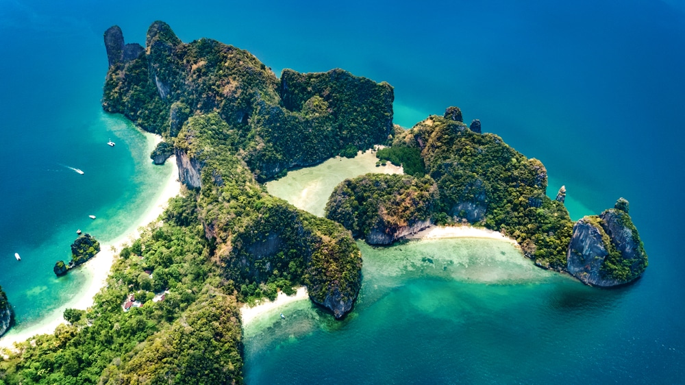 ภาพมุมสูงของเกาะที่เขียวชอุ่มและเต็มไปด้วยโขดหิน มีชายหาดทรายและน้ำทะเลสีฟ้าใส เหมาะสำหรับผู้ที่ต้องการทะเลใกล้กรุงเทพฯ และการพักผ่อนที่เงียบสงบล้อมรอบด้วยมหาสมุทร.