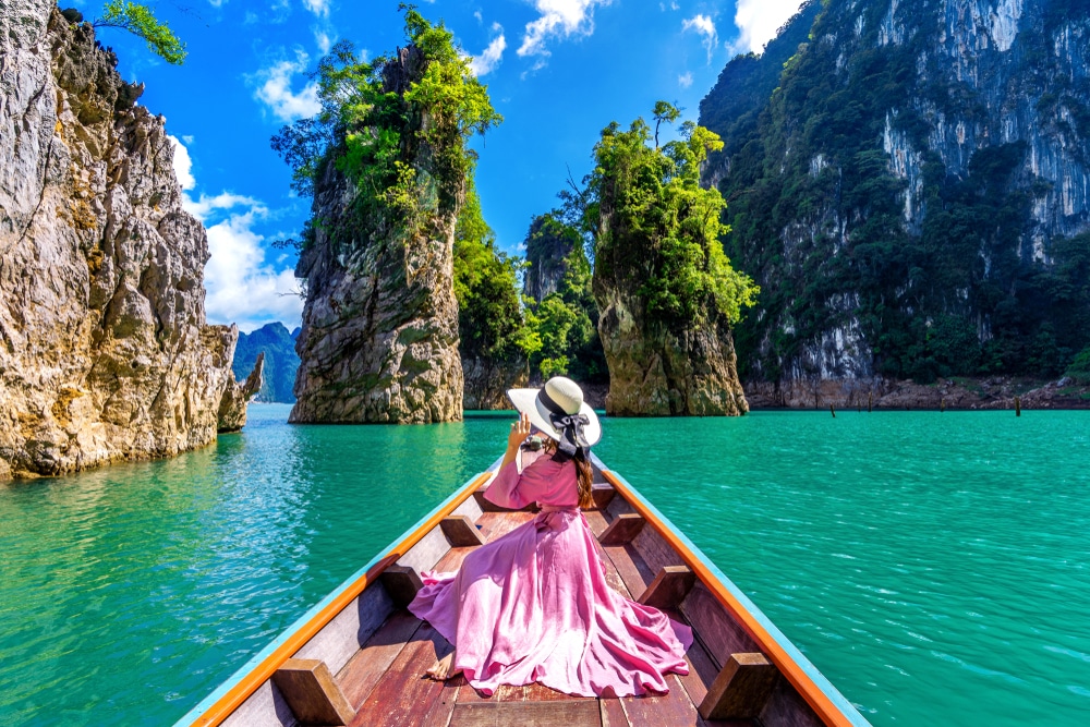 หญิงสาวในชุดเดรสสีชมพูและหมวกปีกกว้างนั่งอยู่ด้านหน้าของเรือไม้ในน้ำสีฟ้าคราม ล้อมรอบด้วยหน้าผาหินสูงชันและพืชพรรณเขียวชอุ่ม—สถานที่ท่องเที่ยวธรรมชาติใกล้กรุงเทพฯ ที่เหมาะสำหรับการหลีกหนีความวุ่นวายเพื่อความสงบ.