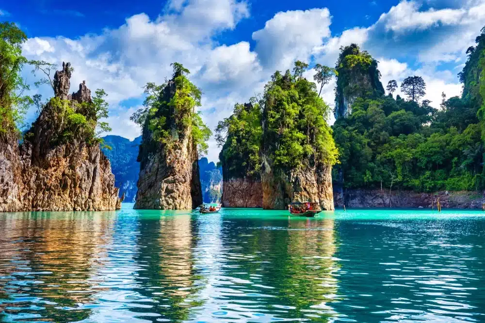 หน้าผาหินปูนสูงตระหง่านปกคลุมด้วยพืชพรรณสีเขียวขจี โผล่ขึ้นจากผืนน้ำสีฟ้าอมเขียวใส มีเรือลำเล็กแล่นลัดเลาะอยู่ระหว่างหน้าผา ท่ามกลางท้องฟ้าสีฟ้าที่มีเมฆบางกระจาย—สถานที่ท่องเที่ยวใกล้กรุงเทพฯ ที่งดงาม เหมาะอย่างยิ่งสำหรับการผจญภัยครั้งต่อไปของคุณ.
