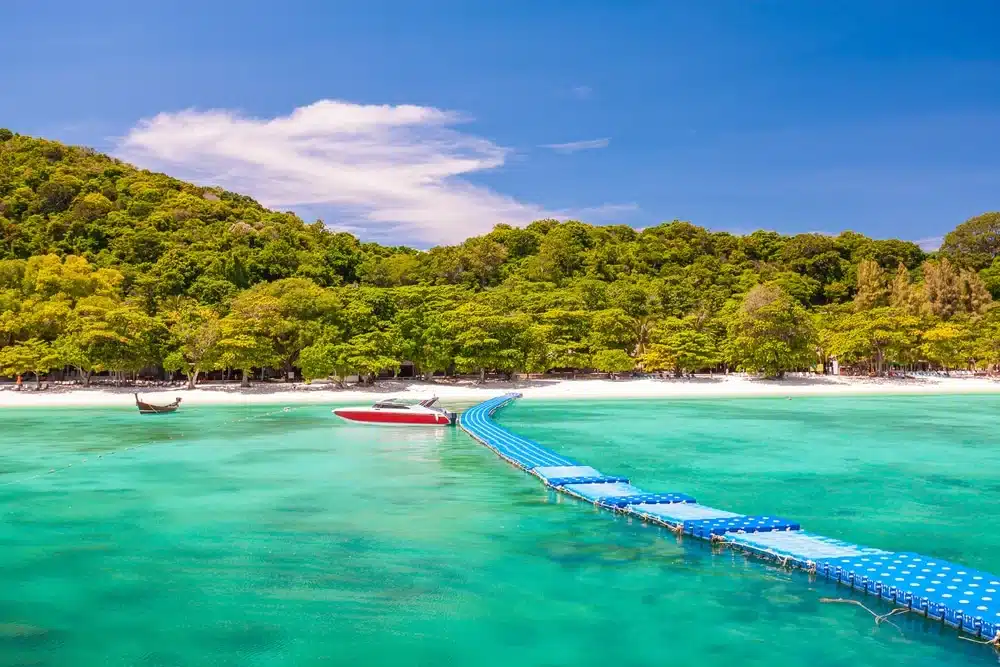 ท่าเรือลอยน้ำสีฟ้ายาวเหยียดจากน้ำทะเลใสสีเทอร์ควอยซ์ไปจนถึงชายหาดทรายขาวที่รายล้อมไปด้วยต้นไม้เขียวชอุ่มหนาแน่น—เหมาะสำหรับผู้ที่ต้องการ ทะเลใกล้กรุงเทพ หรือ ที่เที่ยวธรรมชาติใกล้กรุงเทพ พร้อมเรือเร็วและเรือหางยาวที่จอดอยู่ใต้ท้องฟ้าสีคราม.