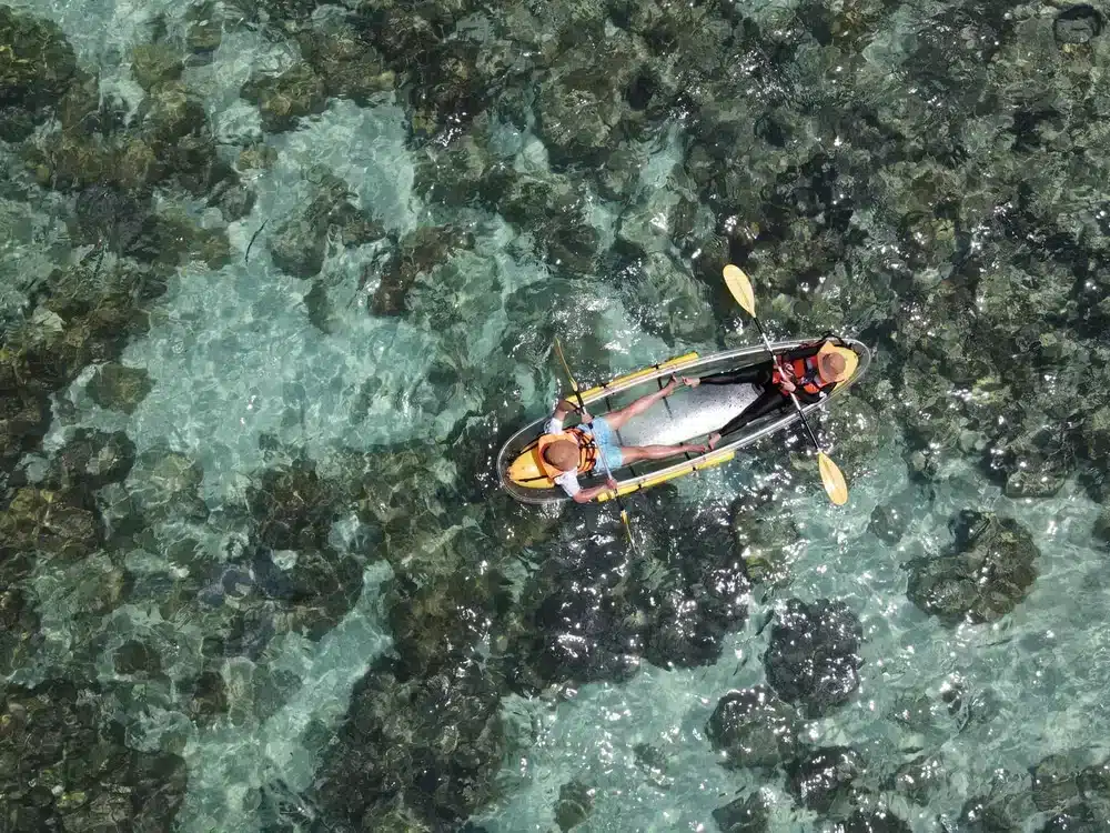 ภาพมุมสูงของสองคนที่กำลังพายเรือแคนูในน้ำใสตื้นซึ่งมองเห็นโขดหินใต้ผิวน้ำได้อย่างชัดเจน—เหมาะสำหรับผู้ที่กำลังมองหากิจกรรมใกล้กรุงเทพฯ เพื่อสัมผัสประสบการณ์ผจญภัยท่ามกลางธรรมชาติในวันหยุด.