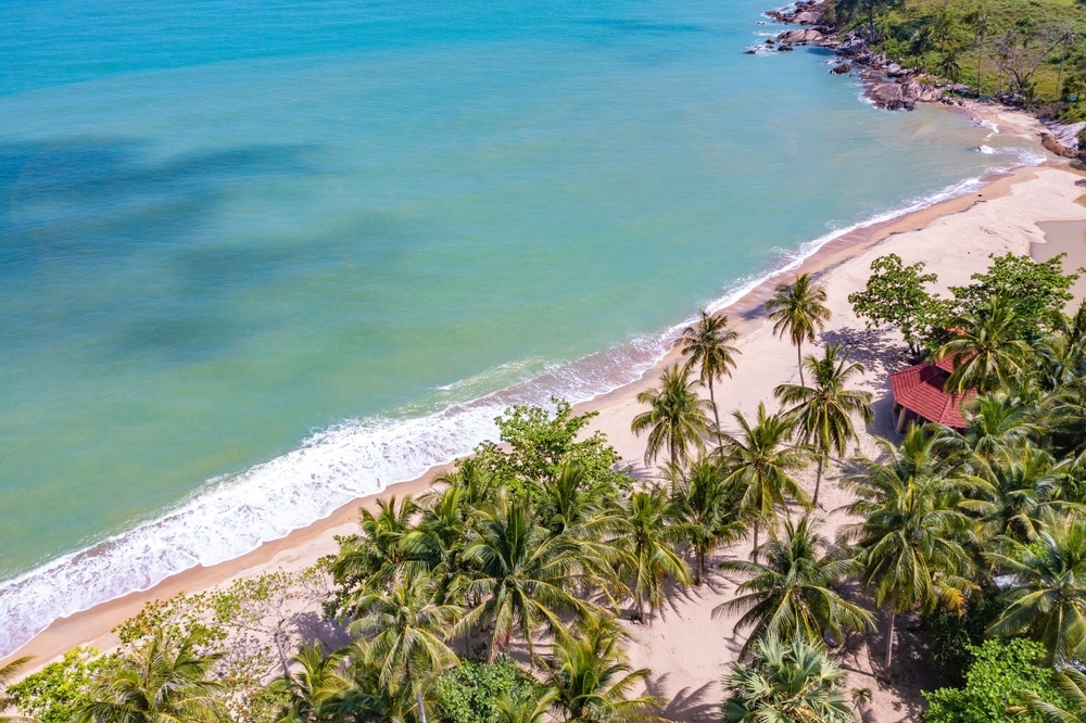 ภาพมุมสูงของชายหาดเขตร้อนที่มีต้นปาล์ม ทรายสีทอง น้ำทะเลสีฟ้าใส และอาคารเล็กๆ ใกล้ชายฝั่ง—เหมาะสำหรับผู้ที่ต้องการ ทะเลใกล้กรุงเทพ หรือ สถานที่ท่องเที่ยวธรรมชาติใกล้กรุงเทพ.