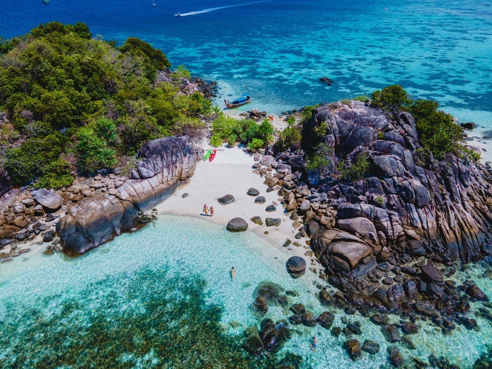 ภาพมุมสูงของเกาะเขตร้อนขนาดเล็กที่มีโขดหินรูปร่างแปลกตา น้ำทะเลใสสีฟ้าเทอร์ควอยซ์ ชายหาดทรายขาวสะอาด และมีผู้คนกับเรือคายัคอยู่ริมฝั่ง—เหมาะอย่างยิ่งสำหรับผู้ที่ต้องการ เที่ยวใกล้กรุงเทพ ในสวรรค์อันเงียบสงบ.