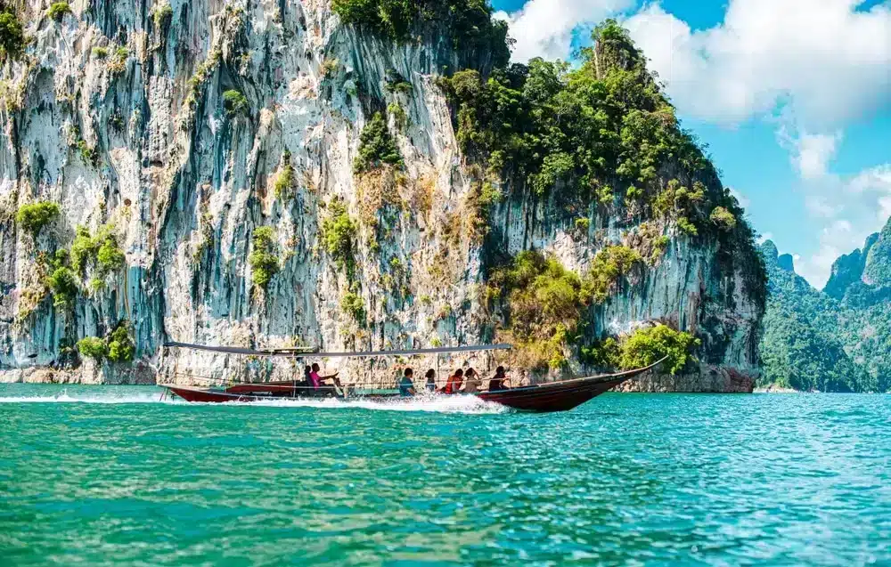 เรือหางยาวไม้บรรทุกผู้โดยสารแล่นผ่านน้ำสีฟ้าครามใกล้หน้าผาหินปูนสูงชันที่ปกคลุมไปด้วยป่าไม้ ท่ามกลางท้องฟ้าสีคราม—เหมาะสำหรับผู้ที่ต้องการสถานที่ท่องเที่ยวธรรมชาติใกล้กรุงเทพฯ และการหลีกหนีความวุ่นวายในเมือง.