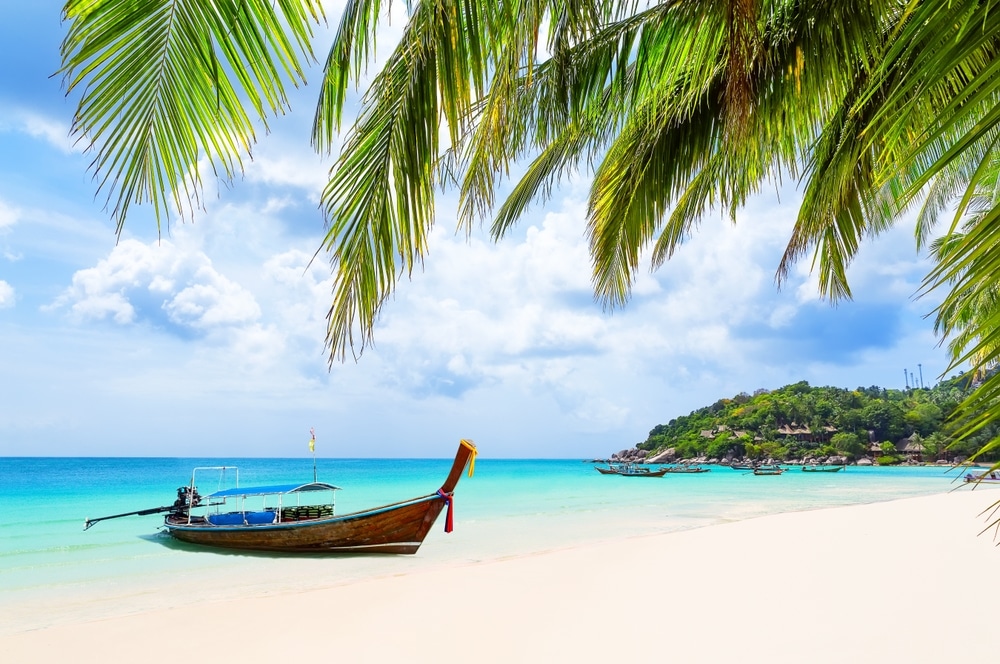 เรือไม้ลำหนึ่งลอยอยู่บนน้ำสีฟ้าใสใกล้ชายหาดทรายที่มีต้นปาล์มอยู่ใต้ท้องฟ้าที่มีเมฆบางส่วนและเนินเขาสีเขียว—เหมาะสำหรับผู้ที่มองหาทะเลใกล้กรุงเทพฯ และสถานที่ท่องเที่ยวธรรมชาติใกล้กรุงเทพฯ.