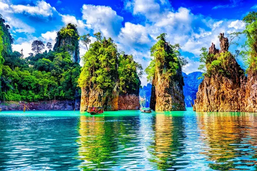 หน้าผาหินปูนสูงใหญ่ปกคลุมด้วยพืชพรรณสีเขียวขจี โอบล้อมน้ำทะเลใสสีฟ้าเทอร์ควอยซ์ ภายใต้ท้องฟ้าสีครามที่มีเมฆบางกระจายอยู่ เป็นภาพที่เหมาะอย่างยิ่งสำหรับสถานที่ท่องเที่ยวธรรมชาติใกล้กรุงเทพฯ สำหรับผู้ที่ต้องการหลีกหนีความวุ่นวายไปสัมผัสธรรมชาติอย่างรวดเร็ว.